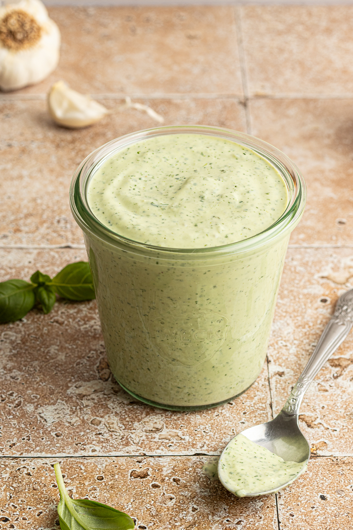 A glass jar filled with creamy green goddess dressing sits on a tile surface, with a spoonful of the dressing resting nearby. Fresh basil leaves and a head of garlic are blurred in the background.