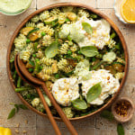 Overhead view of a finished green goddess pasta salad topped with dollops of burrata cheese, pine nuts, and fresh basil. The salad is served in a large wooden bowl surrounded by ingredients like lemon, red pepper flakes, and green herb dressing.