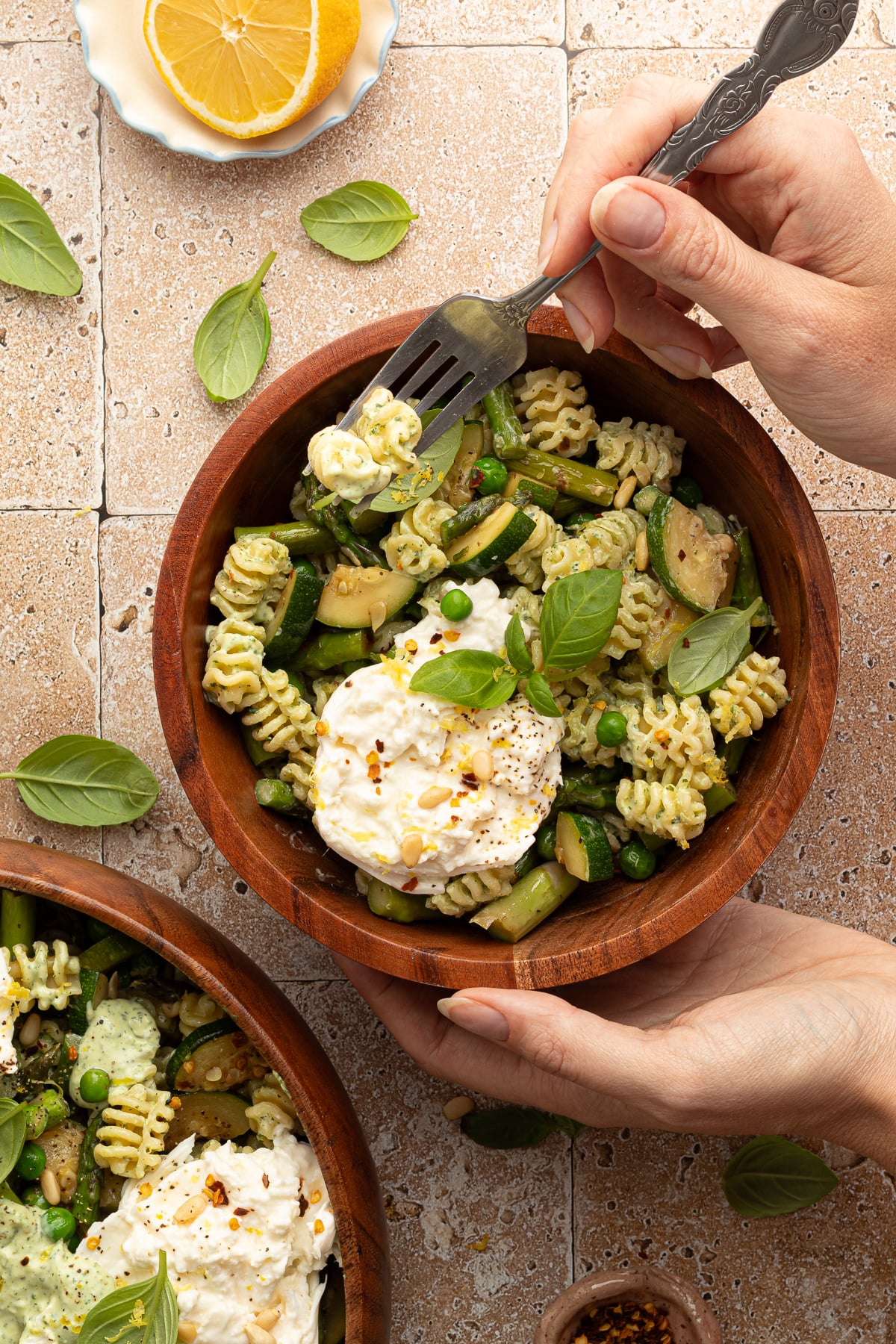 A hand holds a small wooden bowl filled with green goddess pasta salad and a scoop of burrata, while the other hand holds a forkful of pasta. Fresh basil leaves and a cut lemon rest on the tile countertop.