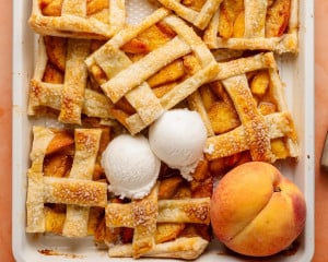 Sliced peach slab pie bars in the baking dish, served with scoops of vanilla ice cream and a whole peach for garnish. The bars are golden, crisp, and ready to enjoy.