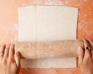 Close-up of puff pastry being rolled out on a floured surface with both hands on the rolling pin. This step is preparing the top lattice crust.