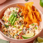 Front-facing view of a bowl of creamy taco pasta salad topped with avocado slices, fresh cilantro, and green onions. A pile of Doritos chips is nestled beside the salad, and a lime wedge is placed in front for garnish.