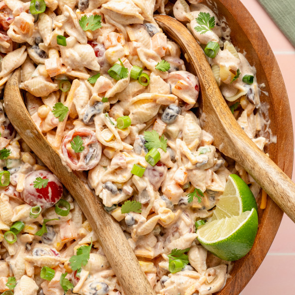 Close-up overhead shot of creamy taco pasta salad in a large wooden bowl with wooden serving spoons. The salad includes shell pasta, cherry tomatoes, corn, black beans, cheese, and is topped with green onions, cilantro, and a lime wedge.