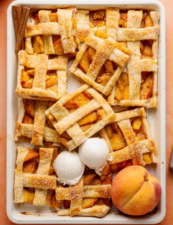 Top-down view of a peach slab pie that has been partially sliced and served, revealing its juicy filling. Three scoops of vanilla ice cream rest on top, and cinnamon sticks and peaches decorate the scene.