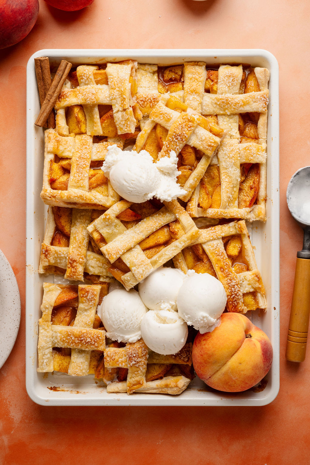 A tray of freshly baked peach slab pie bars with a golden, flaky lattice crust sprinkled with coarse sugar. Several bars are cut and slightly overlapping, topped with scoops of vanilla ice cream melting into the warm pie. A fresh peach and cinnamon sticks are placed beside the tray for garnish. The scene is set on a peach-colored surface with a metal ice cream scoop nearby.