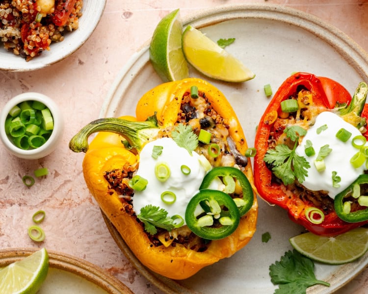 Overhead view of colorful quinoa stuffed bell peppers on a ceramic plate, garnished with sour cream, sliced jalapeños, green onions, and cilantro, with lime wedges on the side.