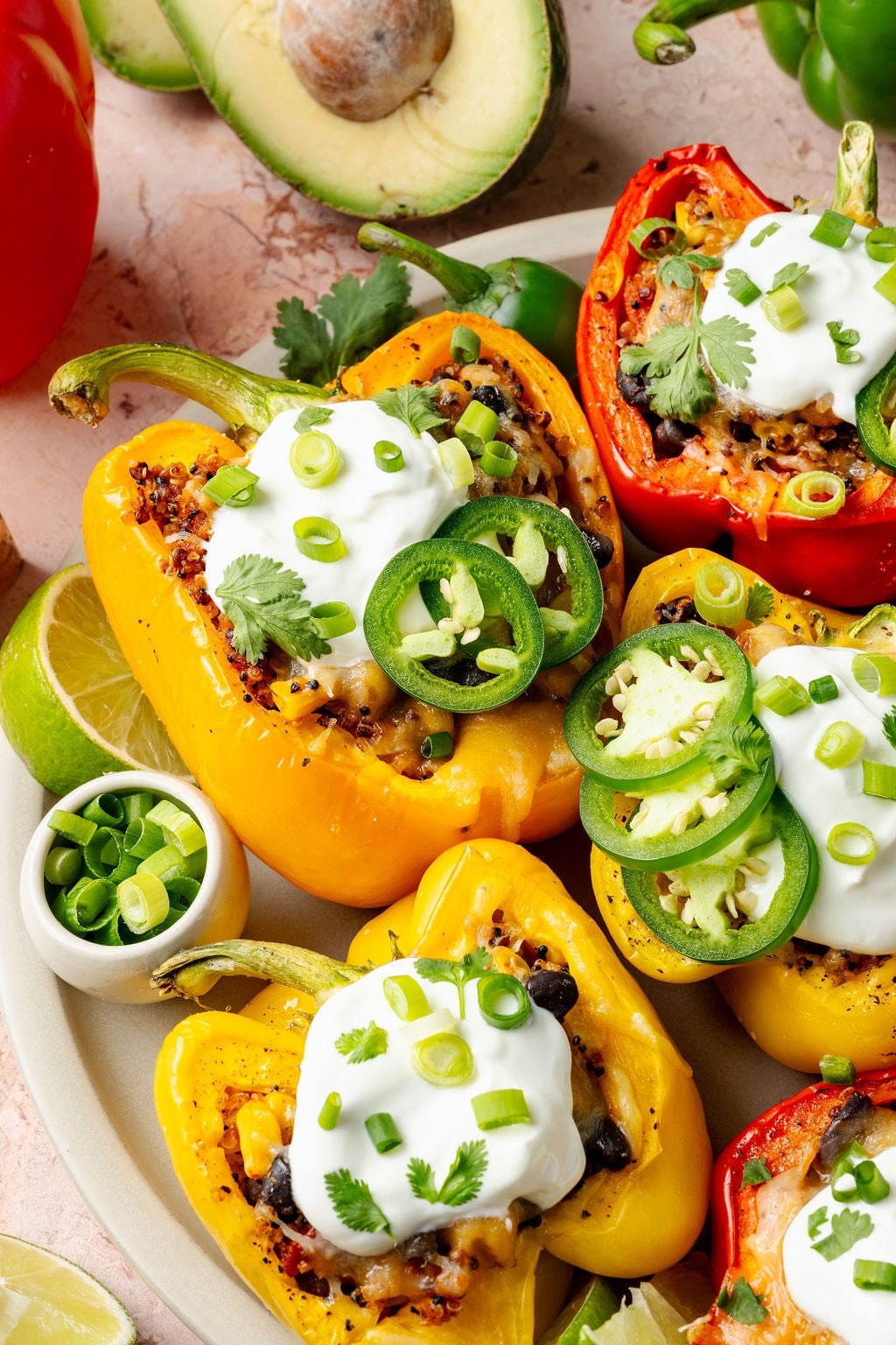 Side angle of a baked red pepper overflowing with a colorful quinoa filling, garnished with a scoop of sour cream and sliced green onions.