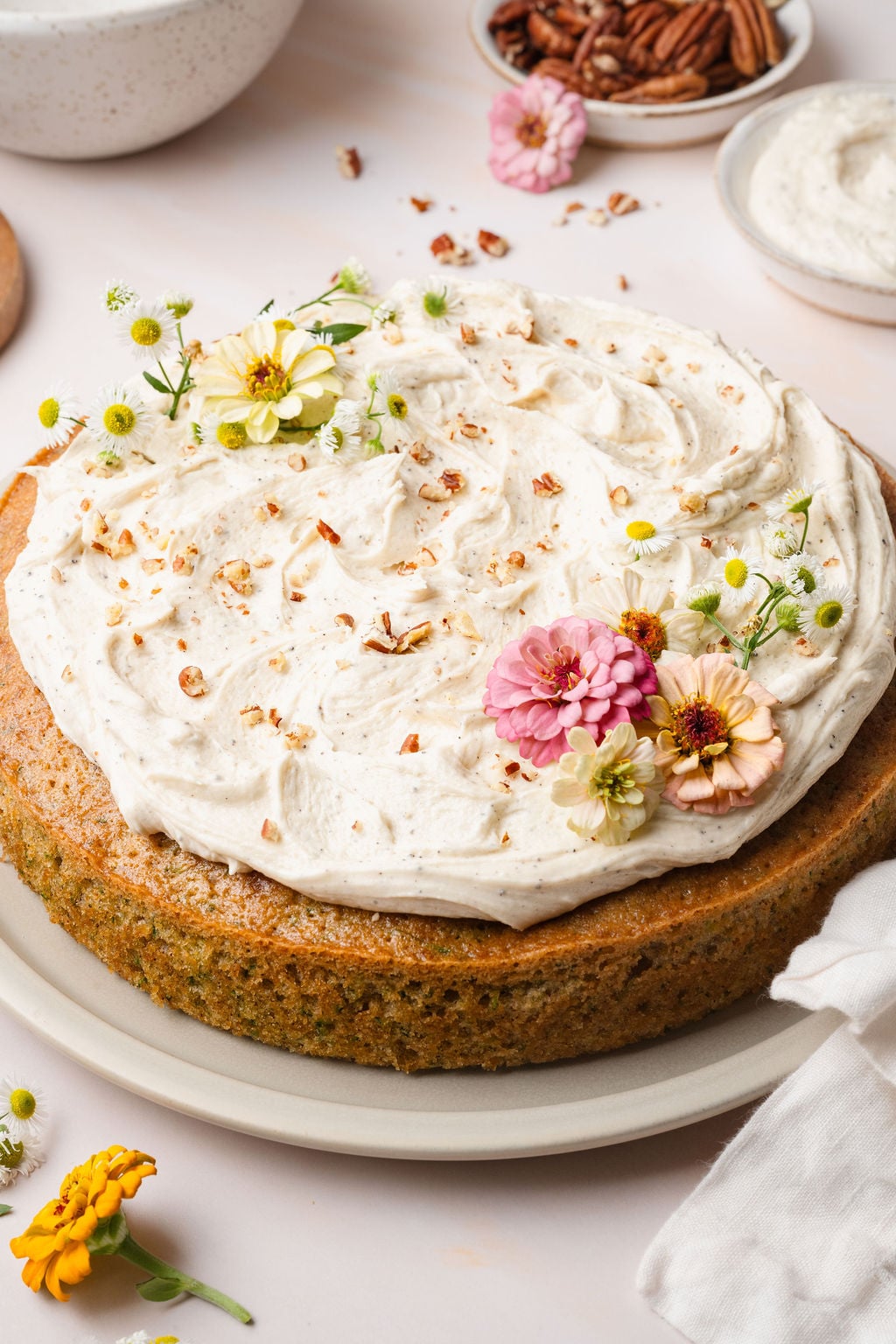 Side view of a whole zucchini cake with thick swirls of vanilla frosting on top, garnished with colorful zinnias and daisies.