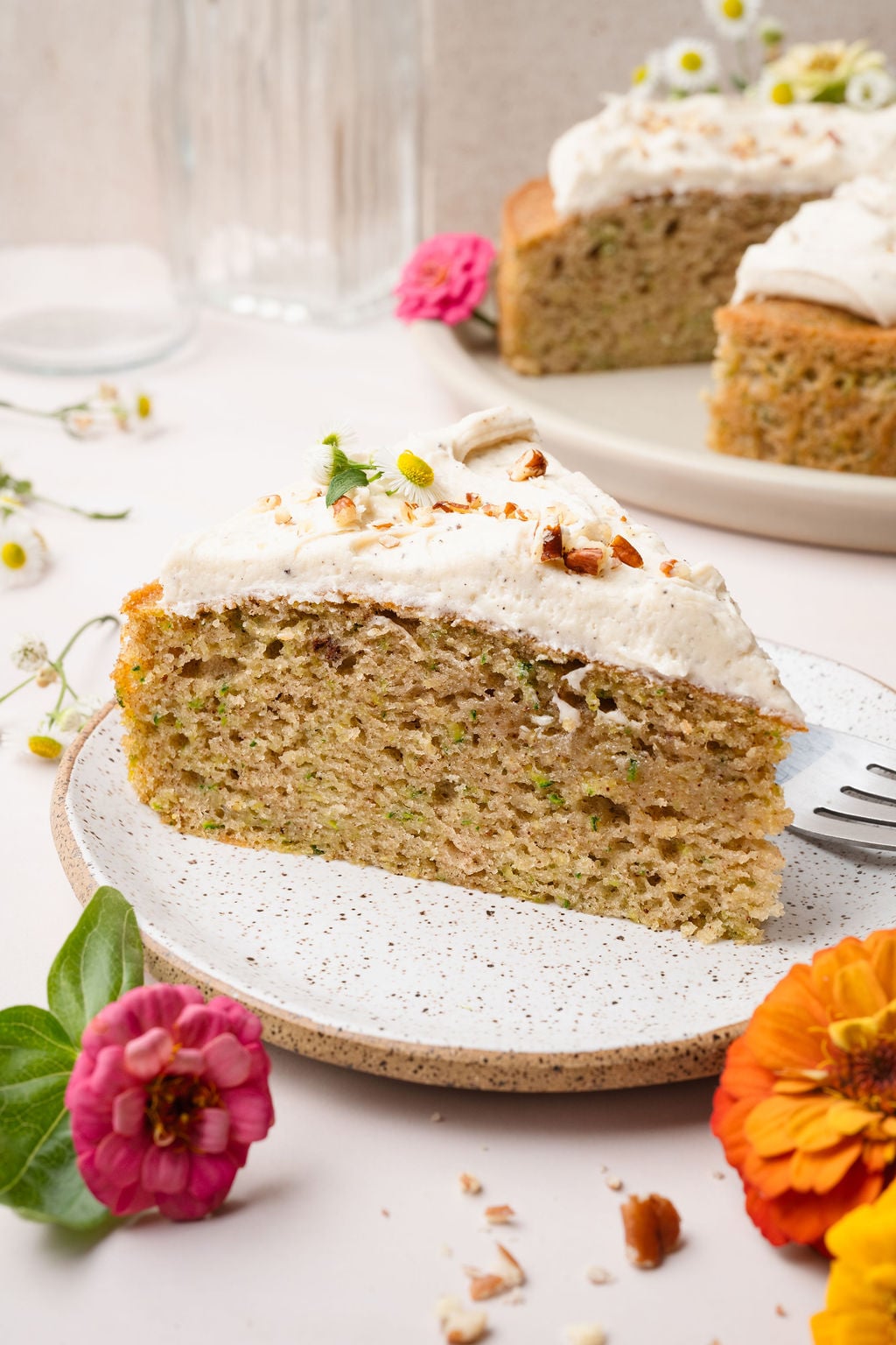 A plated slice of zucchini cake topped with frosting and chopped nuts, with the full cake visible in the background. Surrounded by bright zinnias.