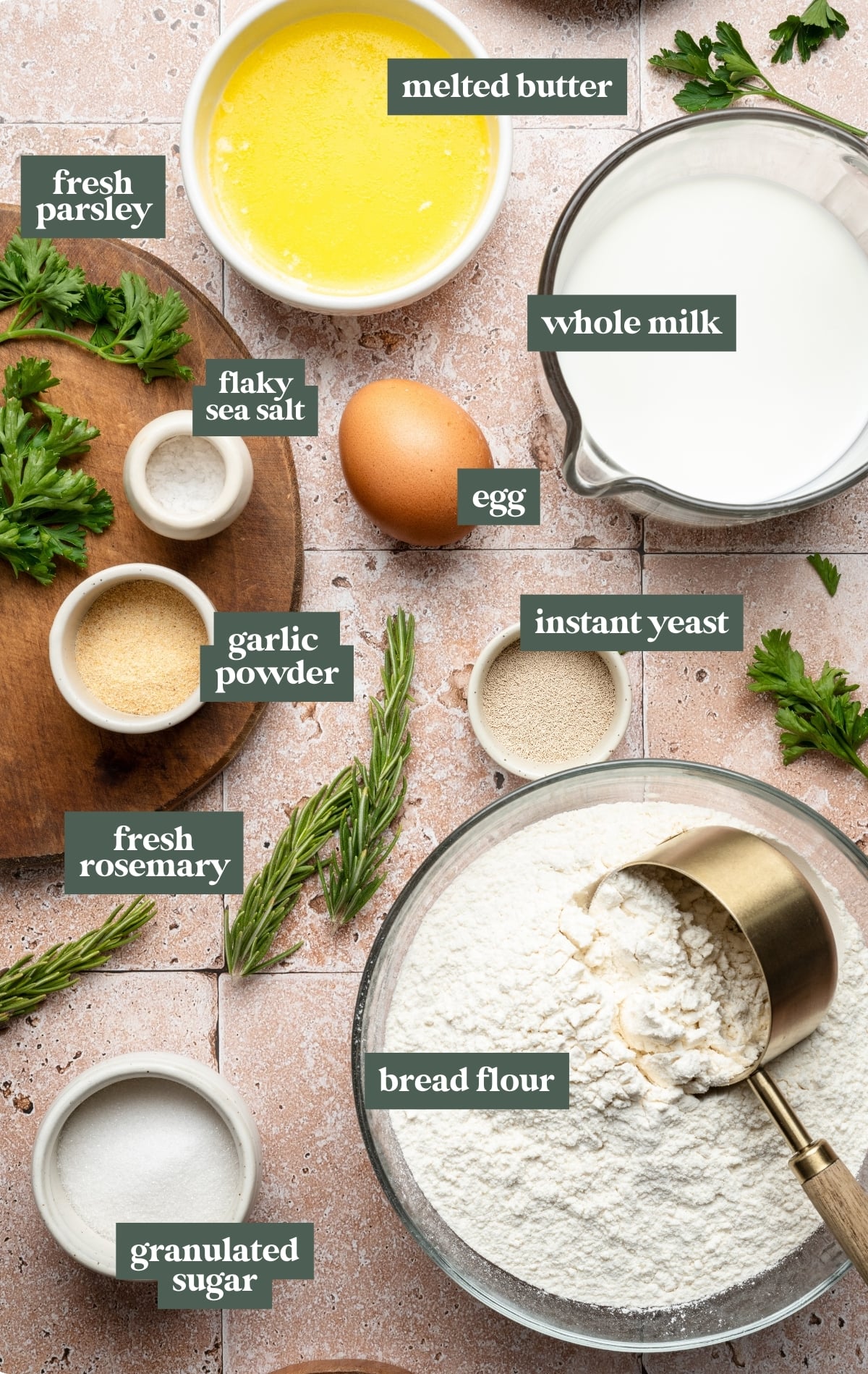 Overhead view of labeled ingredients for garlic rosemary pull-apart rolls, including bread flour, whole milk, egg, melted butter, instant yeast, granulated sugar, garlic powder, flaky sea salt, fresh parsley, and fresh rosemary.