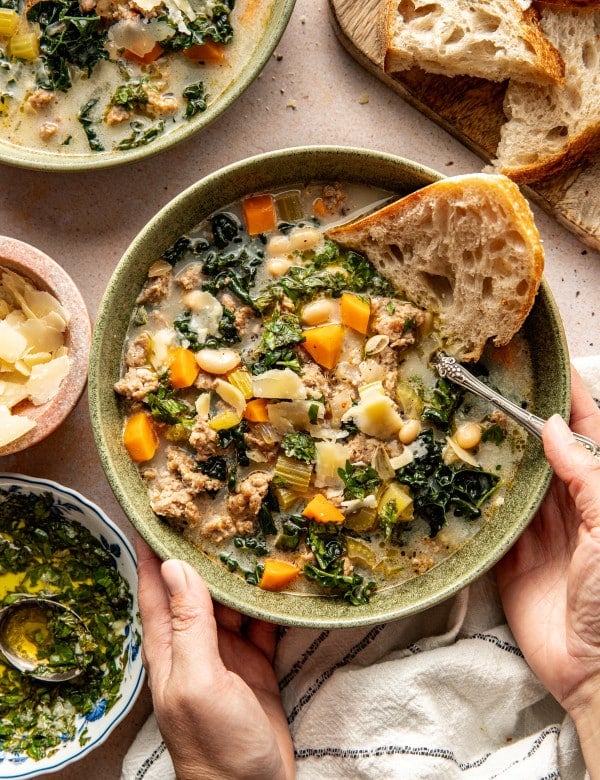 Sausage, kale and white bean soup in bowl with a side of crusty bread and spoon in bowl.