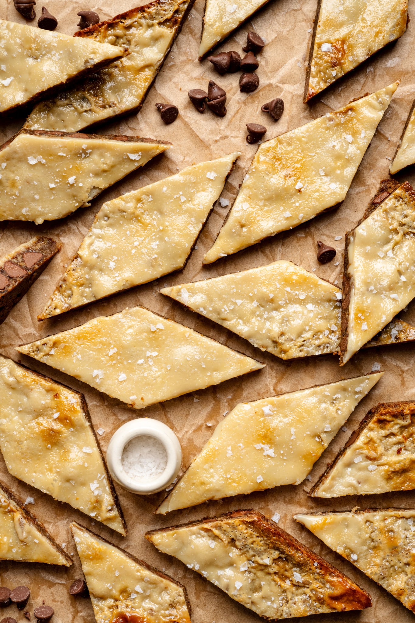 Coffee cookie bars cut into diamond-shapes on parchment paper with small bowl of flaky salt and chocolate chips.