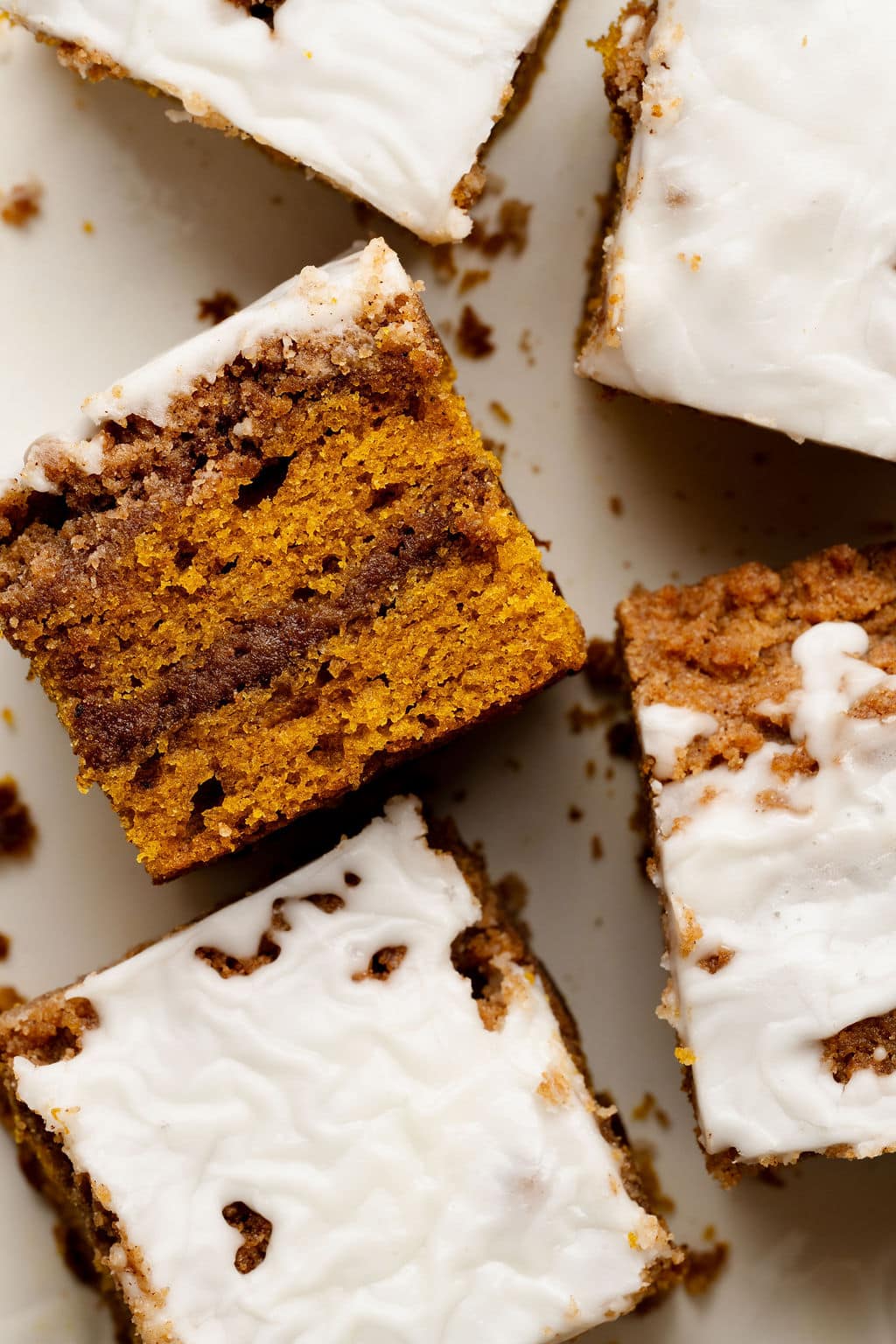 Overhead view of pumpkin coffee cake squares with white glaze, styled on a white plate. The swirl and crumb texture are visible.