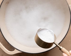 Overhead view of baking soda being added to simmering water in a large pot to prepare a baking soda bath for pretzel bites.