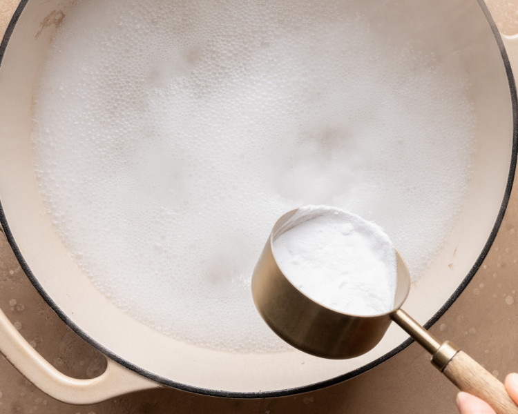 Overhead view of baking soda being added to simmering water in a large pot to prepare a baking soda bath for pretzel bites.