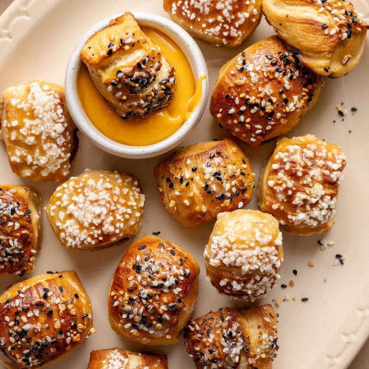 Overhead view of soft, golden-brown pretzel bites on a cream-colored plate, topped with pretzel salt and everything bagel seasoning, with one bite dipped into a bowl of warm cheese sauce in the center.