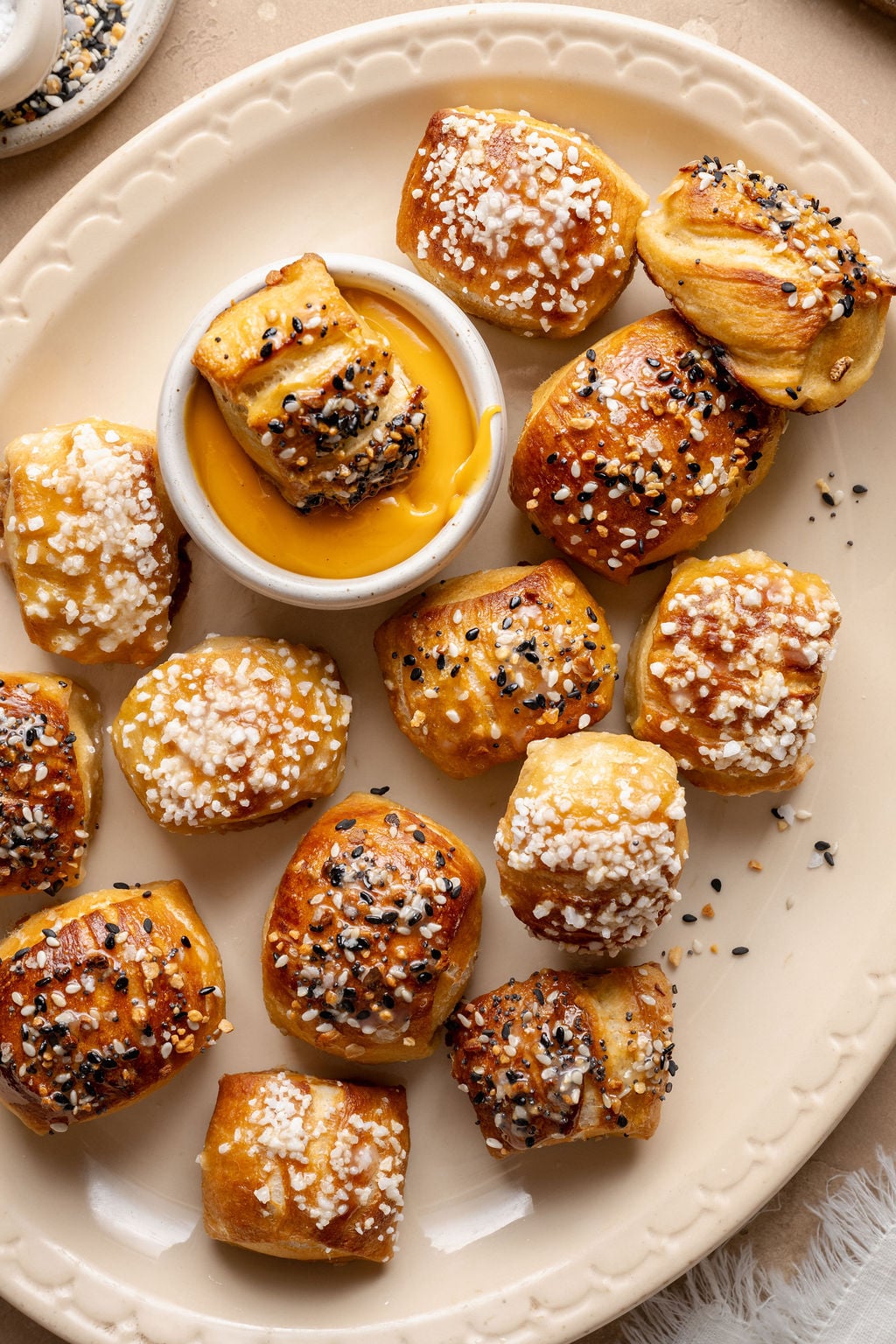 Overhead view of soft, golden-brown pretzel bites on a cream-colored plate, topped with pretzel salt and everything bagel seasoning, with one bite dipped into a bowl of warm cheese sauce in the center.