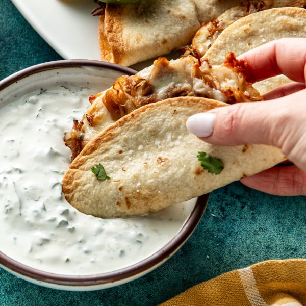 Hand holding pork carnitas taco and dunking it into cilantro crema.