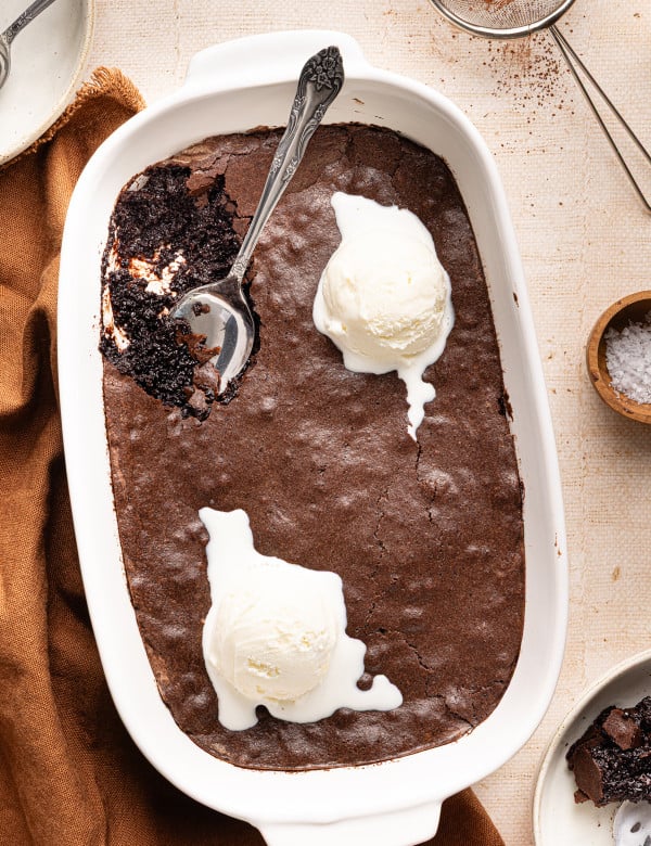Brownie pudding in baking dish with two scoops of vanilla ice cream and spoon in top left corner.