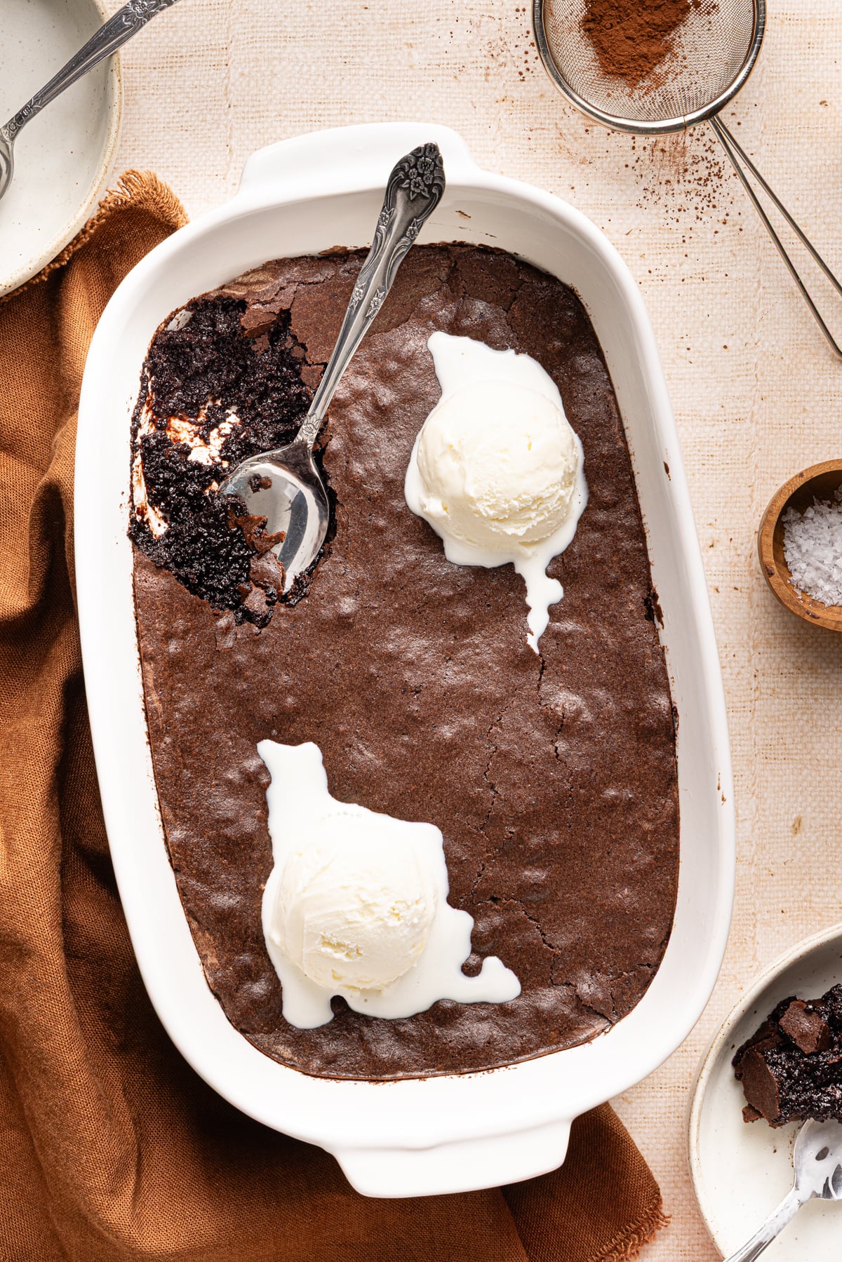 Brownie pudding in baking dish with two scoops of vanilla ice cream and spoon in top left corner.