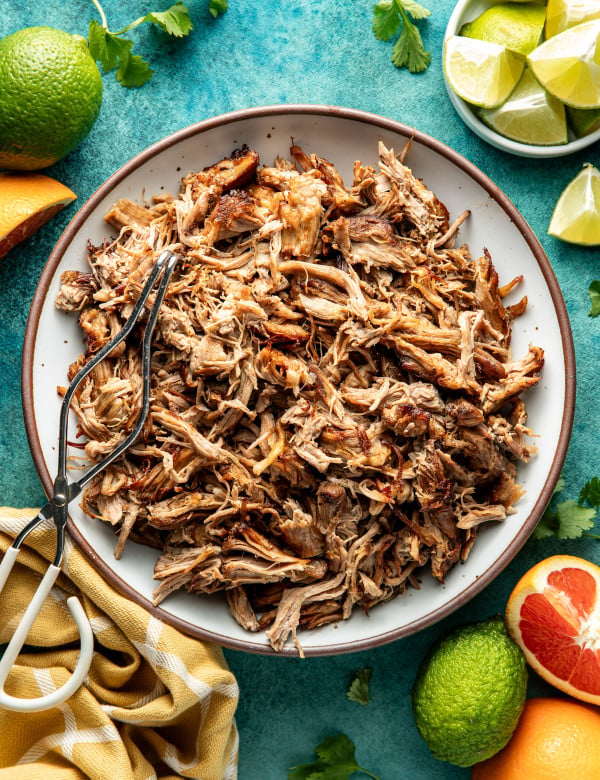 Pork carnitas in white serving bowl with brown rim with metal tongs on top.