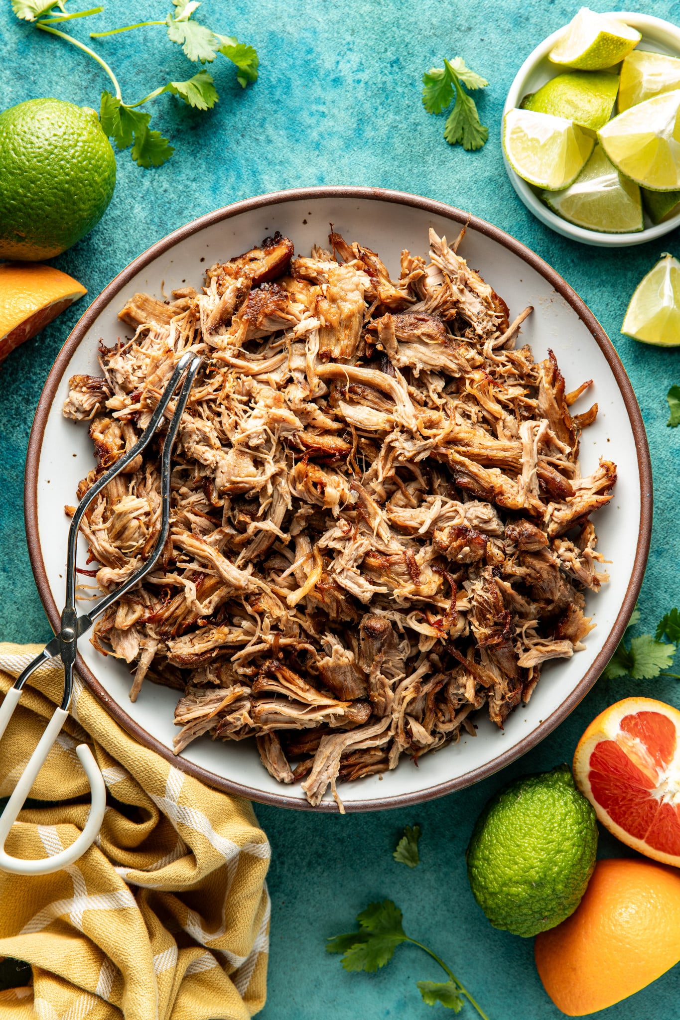 Pork carnitas in white serving bowl with brown rim with metal tongs on top.