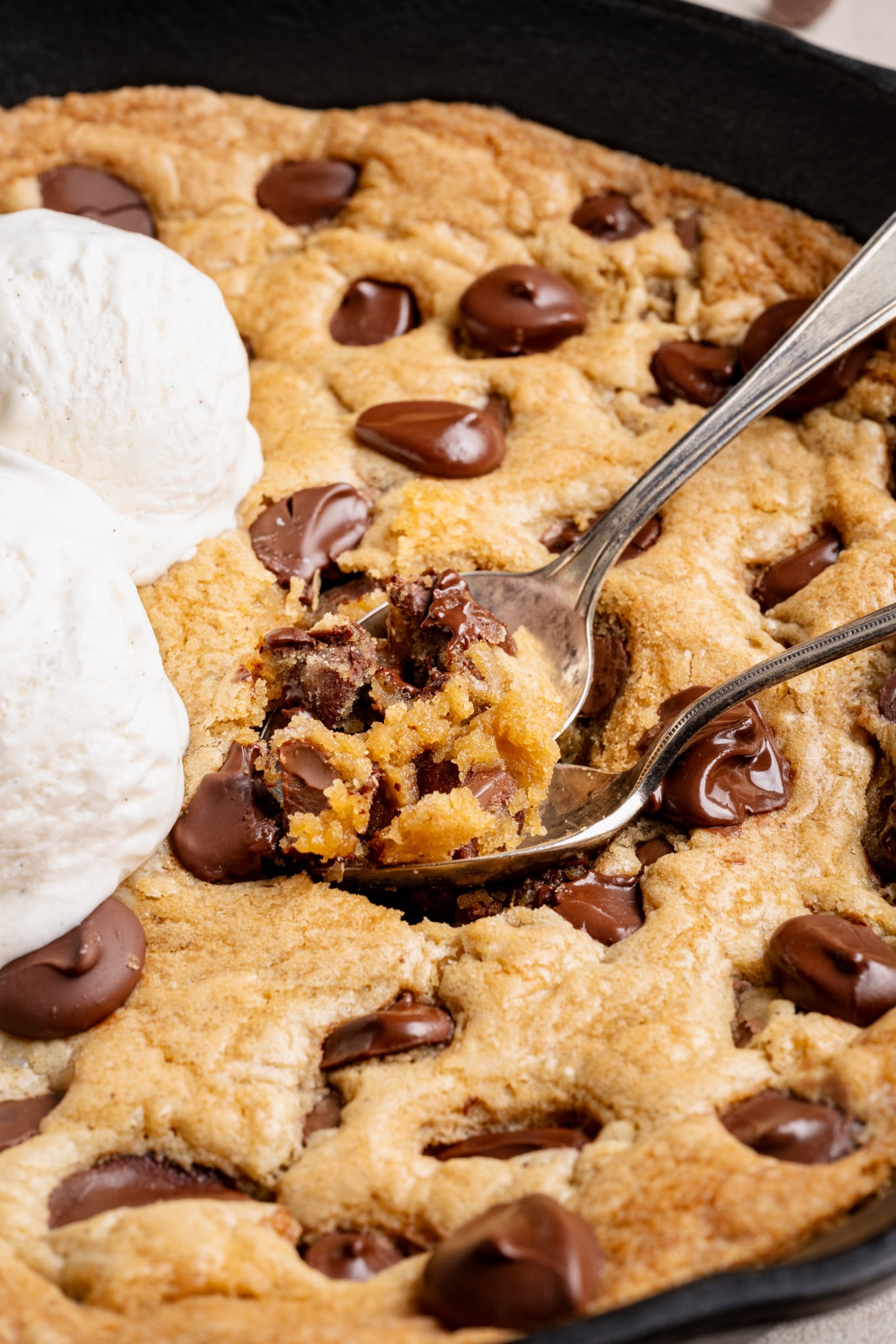 Two spoons digging into a cast iron skillet cookie topped with two scoops of vanilla ice cream.