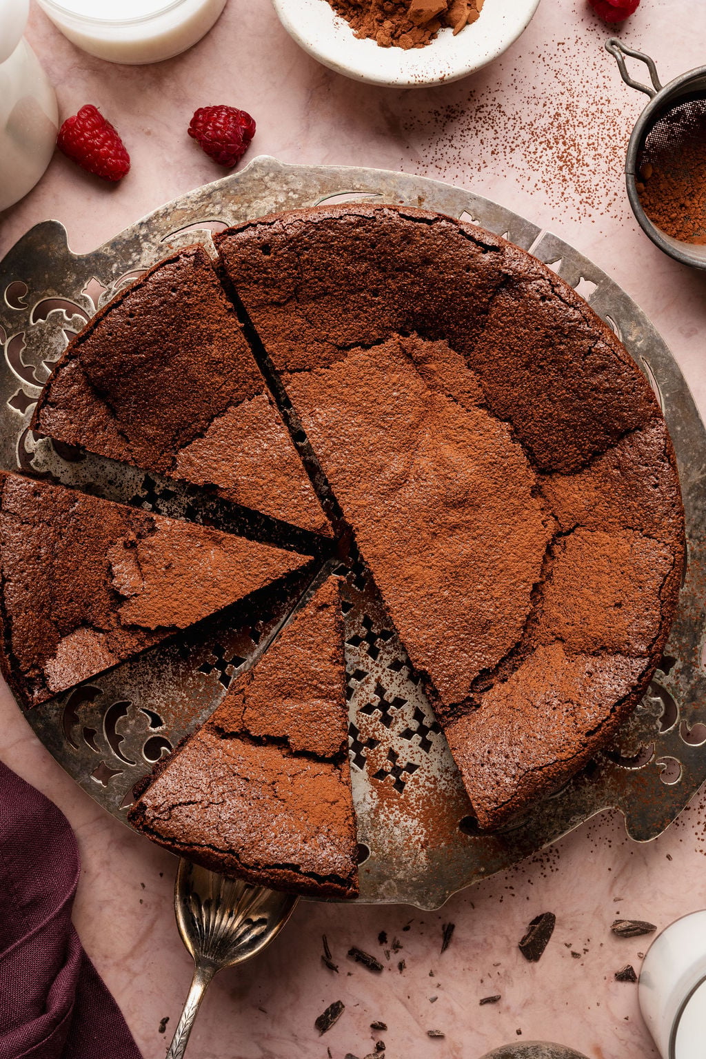 Overhead view of a flourless chocolate cake dusted with cocoa powder, sliced into wedges on a vintage metal cake stand with one slice removed.
