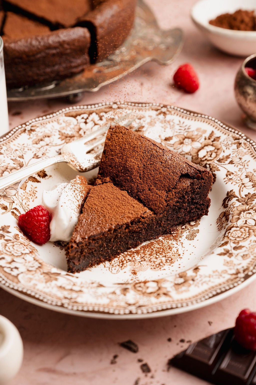 Slice of flourless chocolate cake dusted with cocoa powder on a decorative plate, served with whipped cream and a fresh raspberry, with the remaining cake visible in the background.