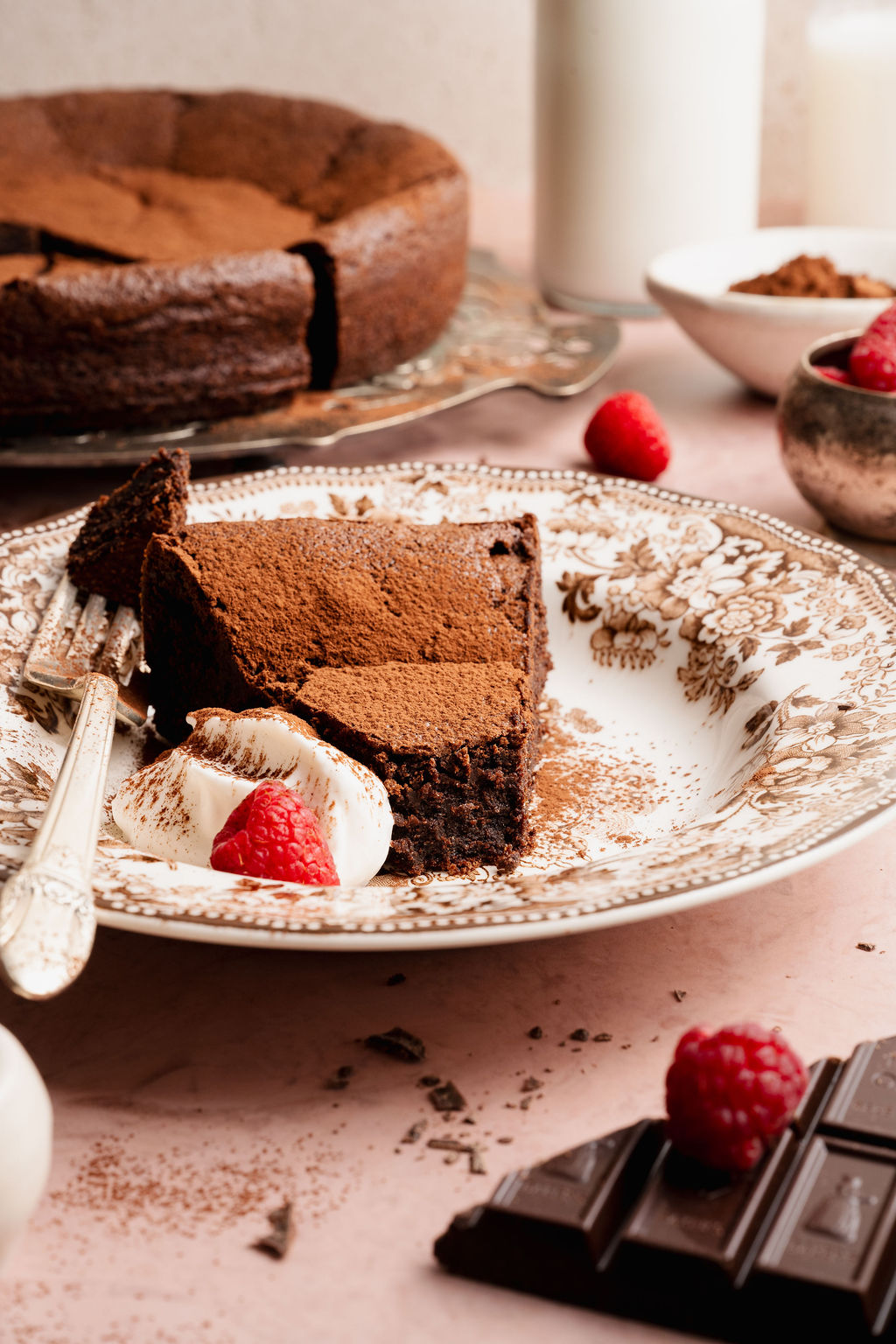 Plated slice of flourless chocolate cake topped with cocoa powder, served with whipped cream and fresh raspberries on a decorative plate.