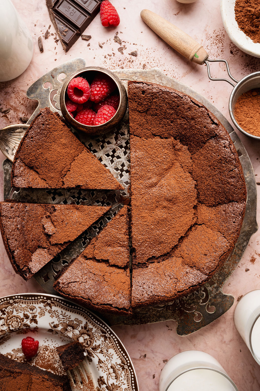 Overhead view of a flourless chocolate cake with multiple slices cut, garnished with cocoa powder and fresh raspberries on a metal cake stand.