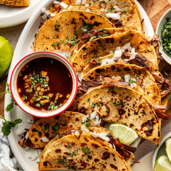 Birria tacos cooked and arranged on serving plate with bowl of consomé on side to dip.