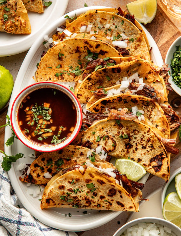 Birria tacos cooked and arranged on serving plate with bowl of consomé on side to dip.