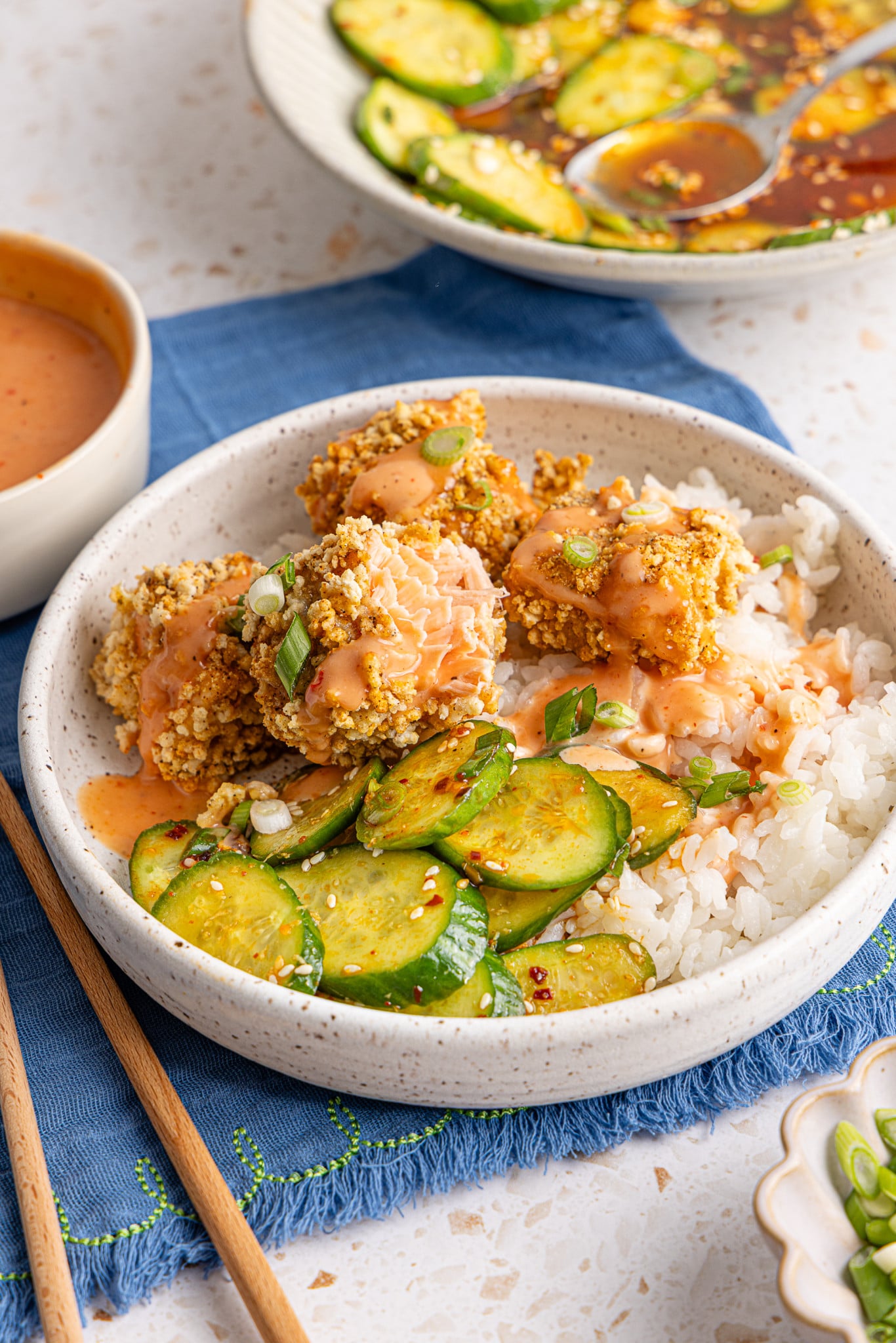 Salmon nuggets served in a bowl with rice and cucumber salad and drizzled with bang bang sauce.