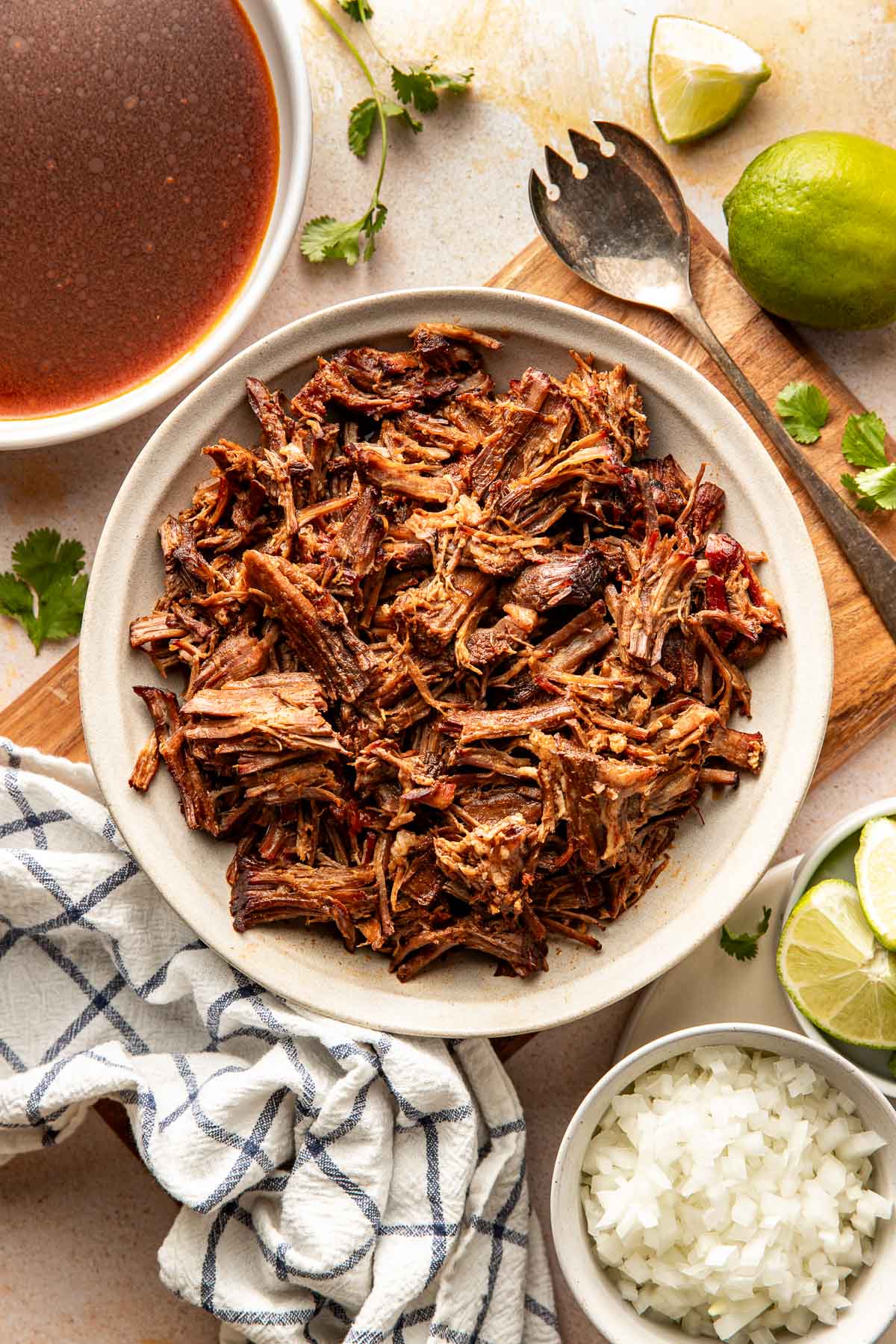 Overhead view of a bowl filled with shredded birria beef, with a bowl of reddish birria broth, lime wedges, cilantro, and diced onions arranged around it.