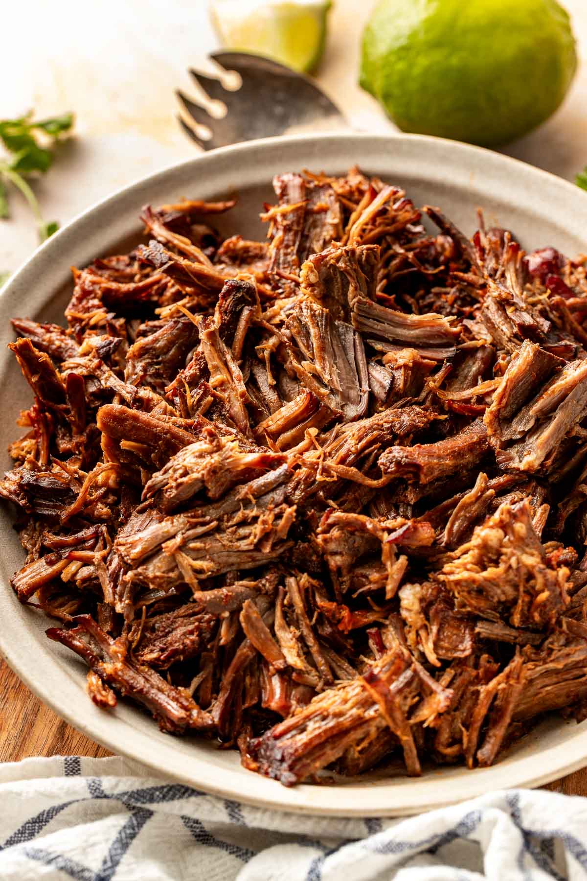 Angled close-up of shredded birria beef in a bowl with visible strands of meat and lightly crisped edges, styled with lime and cilantro in the background.