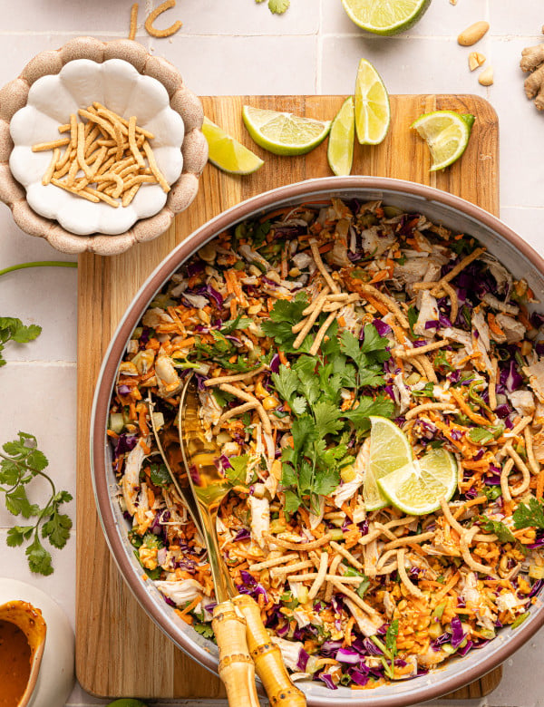 Thai chicken salad with peanut dressing in large bowl garnished with chow mein noodles and lime wedges.