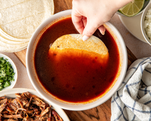 Dipping a white corn tortilla into warm consomé before adding it to the hot skillet.