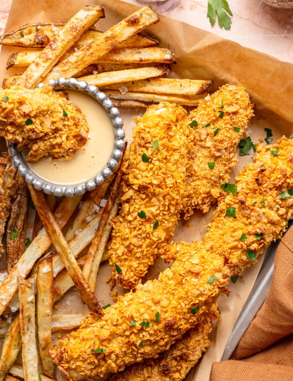 Tray of cornflake chicken tenders with fries and honey mustard dipping sauce with one tender being dunked into the dipping sauce.