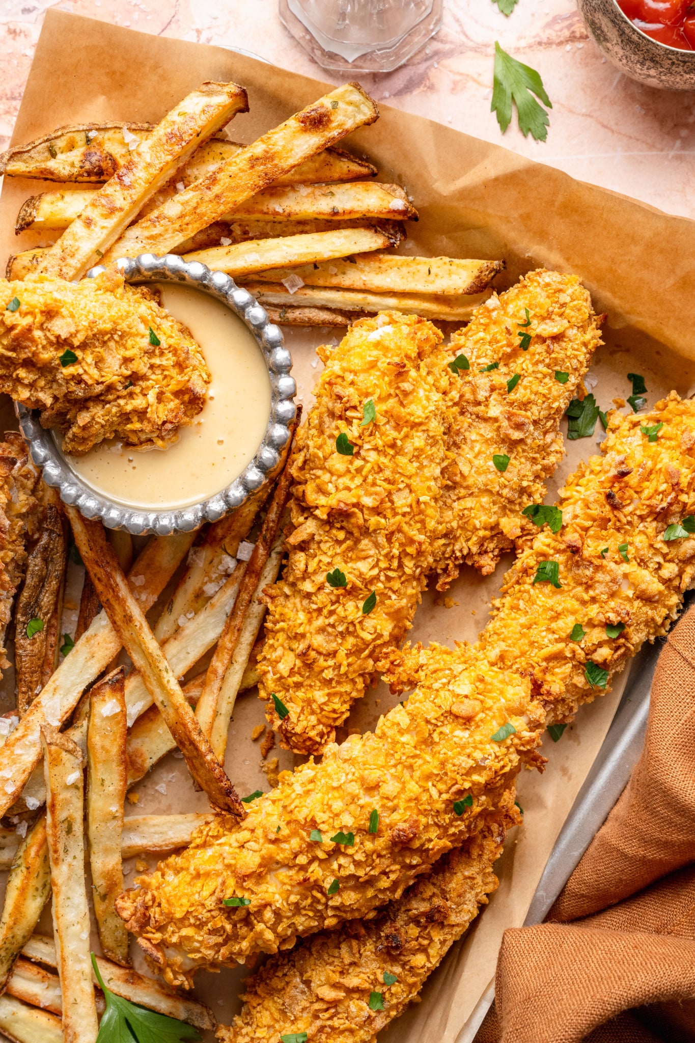 Tray of cornflake chicken tenders with fries and honey mustard dipping sauce with one tender being dunked into the dipping sauce.