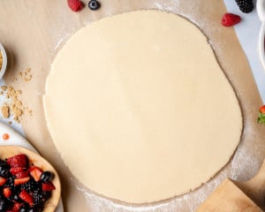 Overhead view of galette dough rolled into a large round on parchment paper, lightly floured surface, with berries and baking tools nearby.