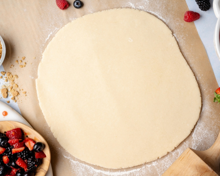 Overhead view of galette dough rolled into a large round on parchment paper, lightly floured surface, with berries and baking tools nearby.