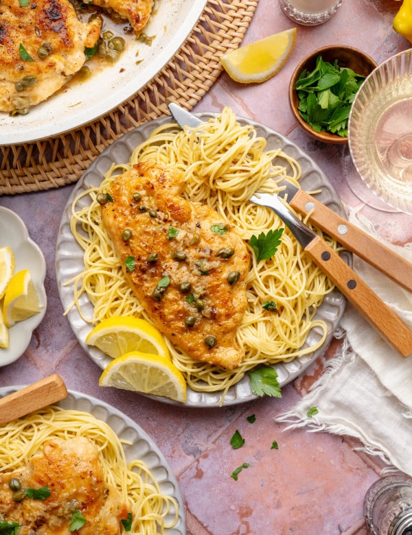Chicken piccata served on top of spaghetti pasta on a plate with fork and knife.
