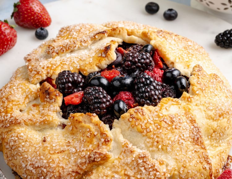 Golden, rustic mixed berry galette with a flaky, sugar-sprinkled crust folded around a juicy filling of blackberries, blueberries, and sliced strawberries, served on a white plate. Fresh berries are scattered around the plate, with a small colander of berries and bottles of milk softly blurred in the background, creating a bright, fresh kitchen scene.