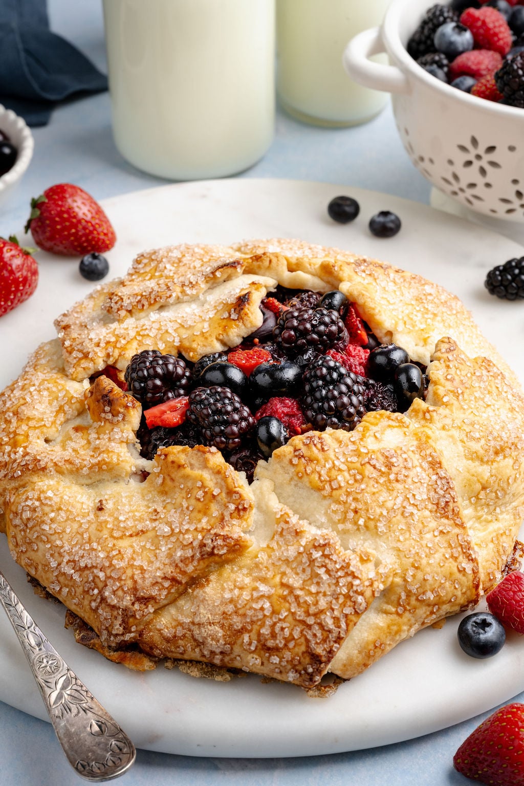 Golden, rustic mixed berry galette with a flaky, sugar-sprinkled crust folded around a juicy filling of blackberries, blueberries, and sliced strawberries, served on a white plate. Fresh berries are scattered around the plate, with a small colander of berries and bottles of milk softly blurred in the background, creating a bright, fresh kitchen scene.