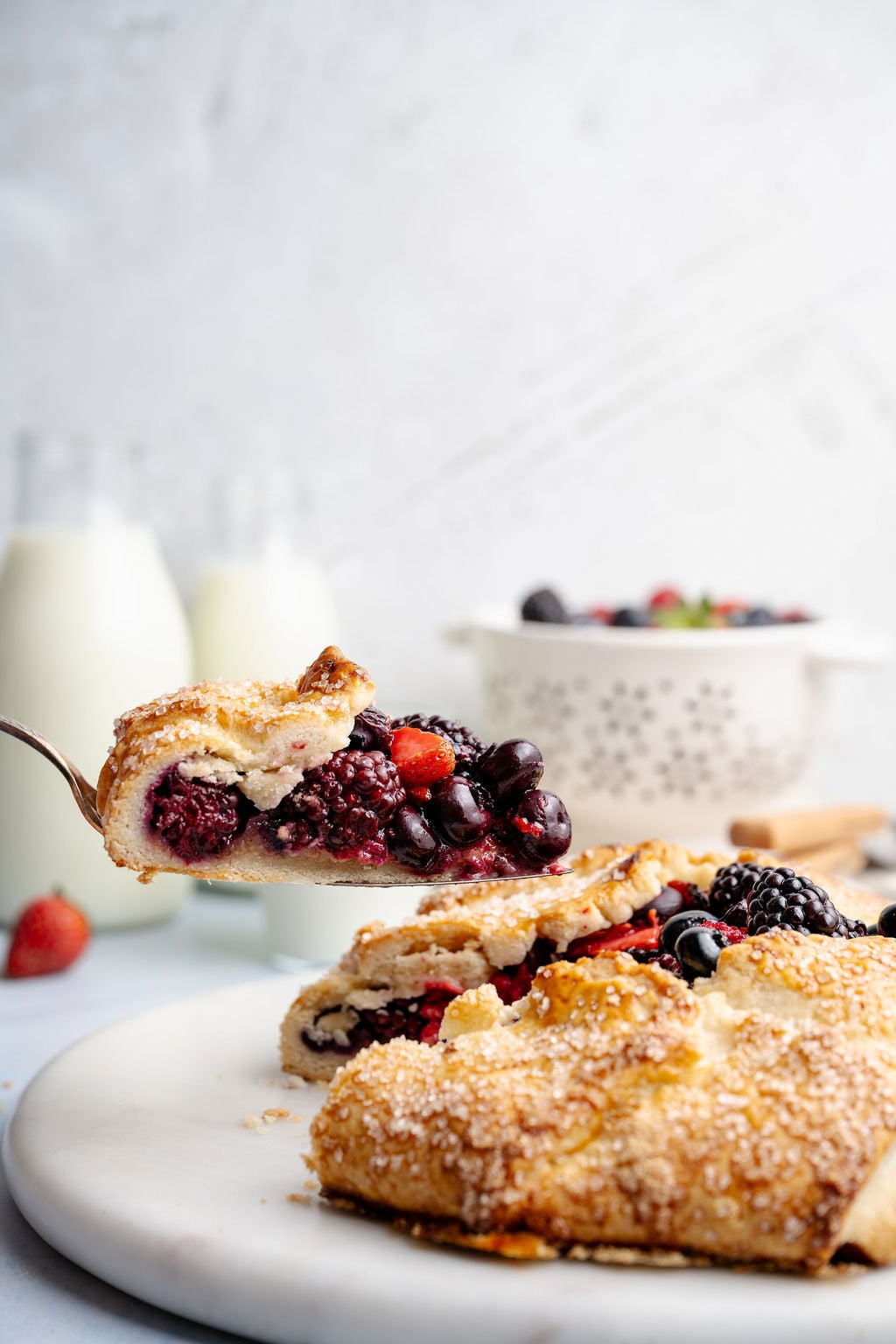 Close-up of a slice of mixed berry galette lifted on a spoon, showing juicy blackberries, blueberries, and strawberries inside a flaky, golden crust with coarse sugar.