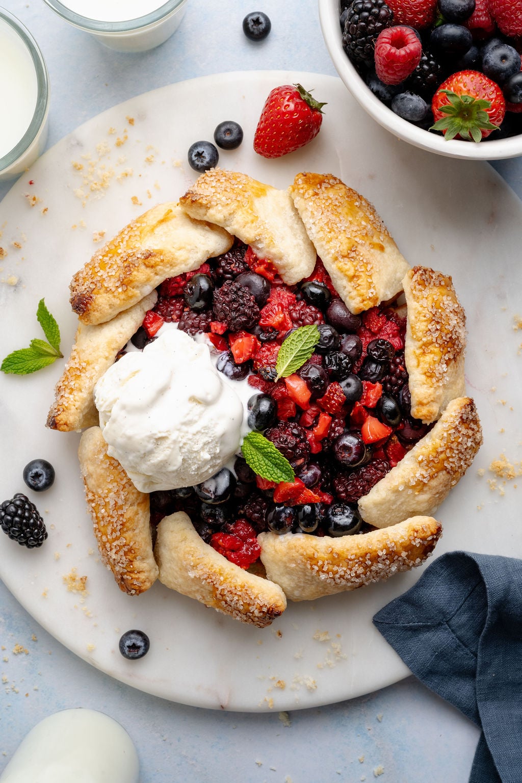 Overhead view of a mixed berry galette topped with a scoop of vanilla ice cream, garnished with fresh mint and surrounded by scattered berries and crumbs.