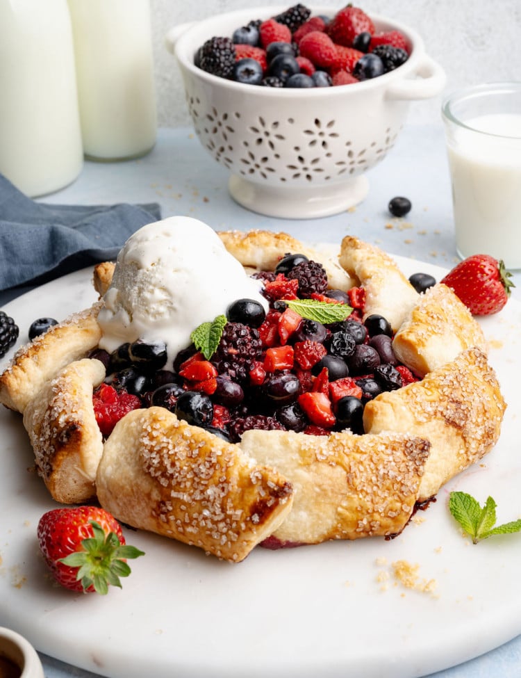 Close-up angled view of a baked mixed berry galette with a scoop of melting vanilla ice cream, highlighting the golden, flaky crust and vibrant berry filling.