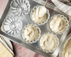 Close-up of a glass dessert cup filled with white chocolate mousse topped with whipped cream, fresh raspberries, and white chocolate curls, with more mousse cups softly blurred in the background.