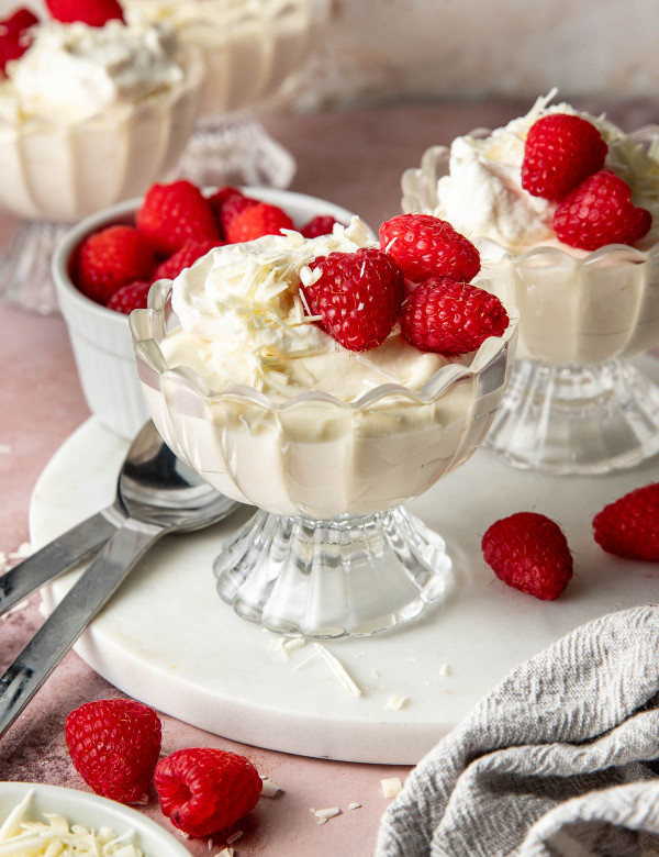 Angled hero shot of white chocolate mousse served in scalloped glass dessert cups topped with whipped cream, raspberries, and white chocolate shavings. Fresh raspberries, dessert spoons, and a linen towel are styled around the desserts on a pink tabletop.